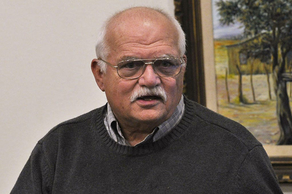 Fred Bailey during a lecture An older man wearing glasses and a dark gray sweater stands indoors in front of a painting.