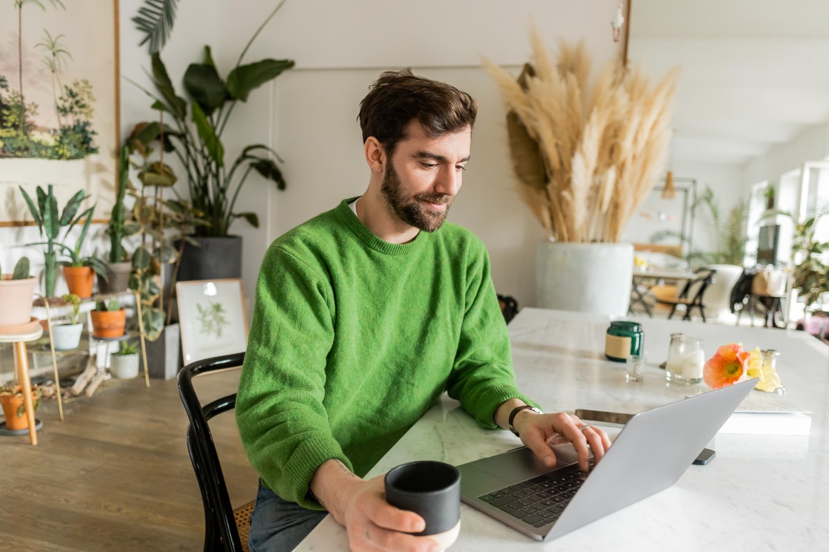 Smiling man in sweater works on a laptop in a room decorated with plants.