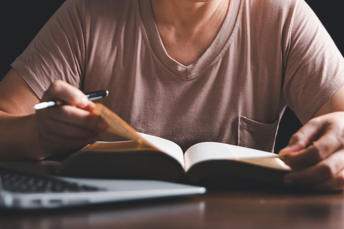 Person studying at a table, reading an open book beside a laptop and a coffee cup.