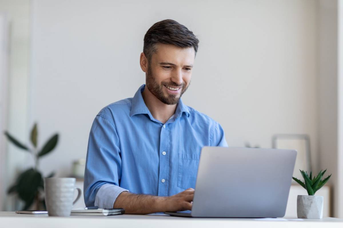 Smiling man in a blue shirt working on a laptop in a modern home office environment with coffee mug and plant on desk