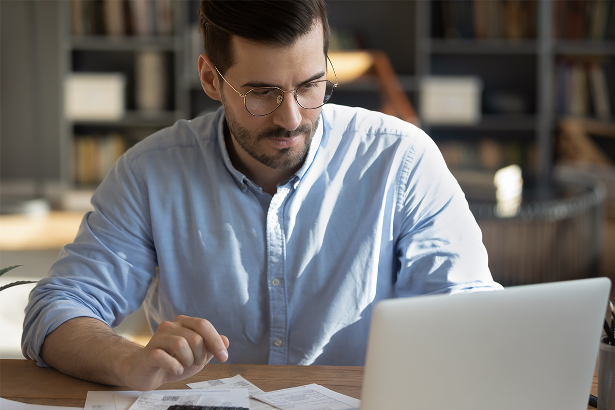 Man wearing glasses working on a computer with papers spread out in front of him, focused on reviewing documents in an office setting. 