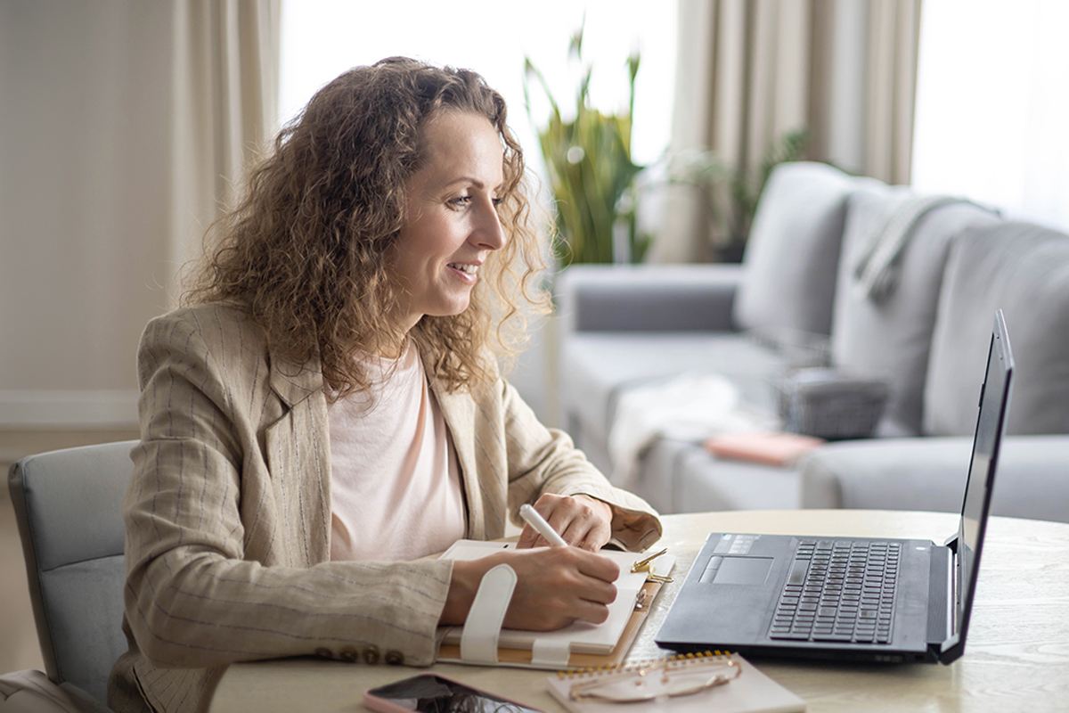 Woman sitting at a desk using a computer while speaking and taking notes in a notepad during an online meeting.