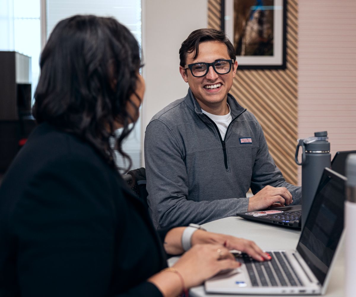 A man and a woman sitting at a desk with laptops in from of them talking to each other.