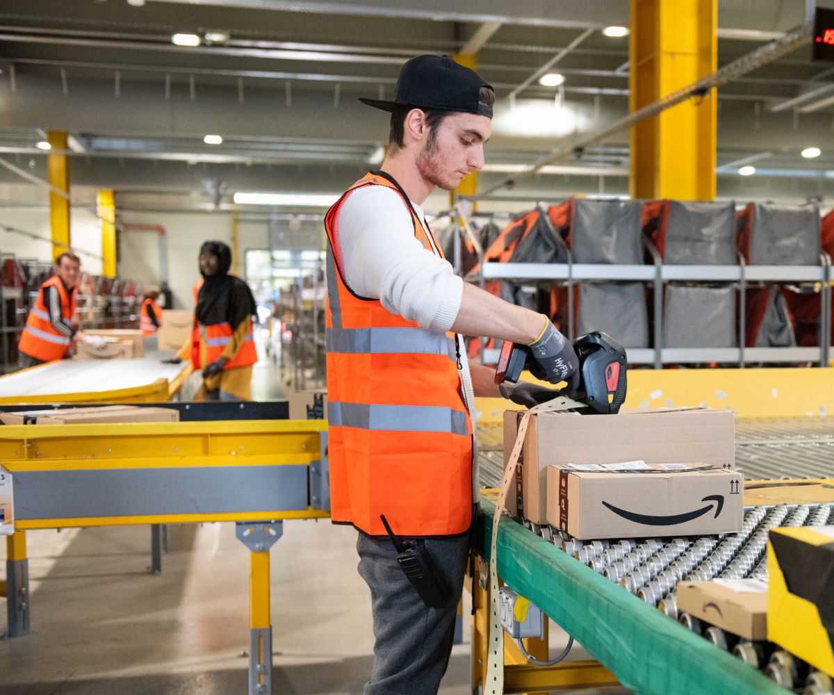 A worker wearing a construction safety vest focusing scanning a package with several additional coworkers in the background.