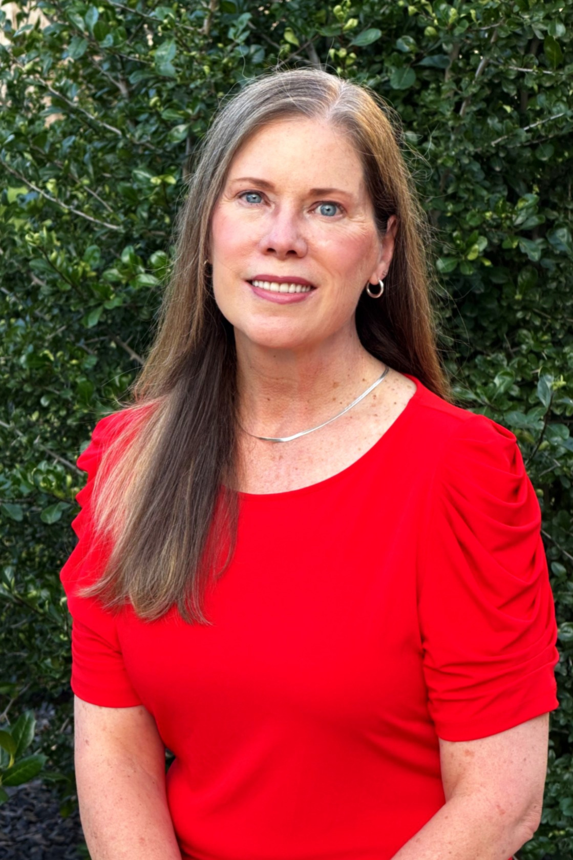 Woman with long brown hair wearing a red top seated in front of greenery