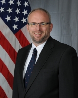Headshot of a man in a blue suit with a bluish-gray background and an American flag