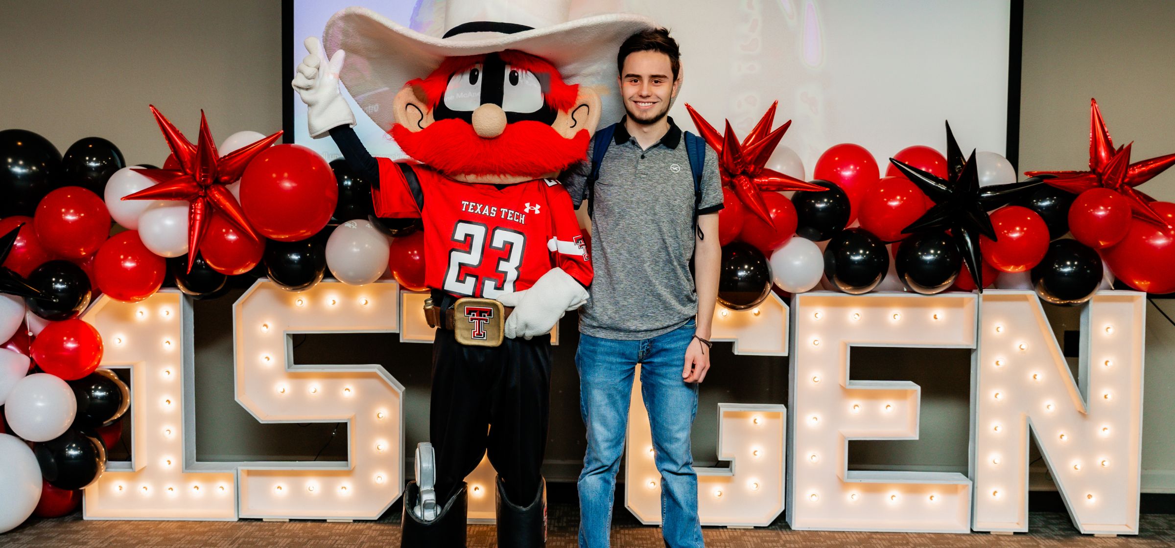 Student next to the mascot, Raider Red standing in front of a sign that says 1st Gen.