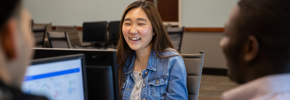 Close up of student at computer, smiling to two companions who are blurred.
