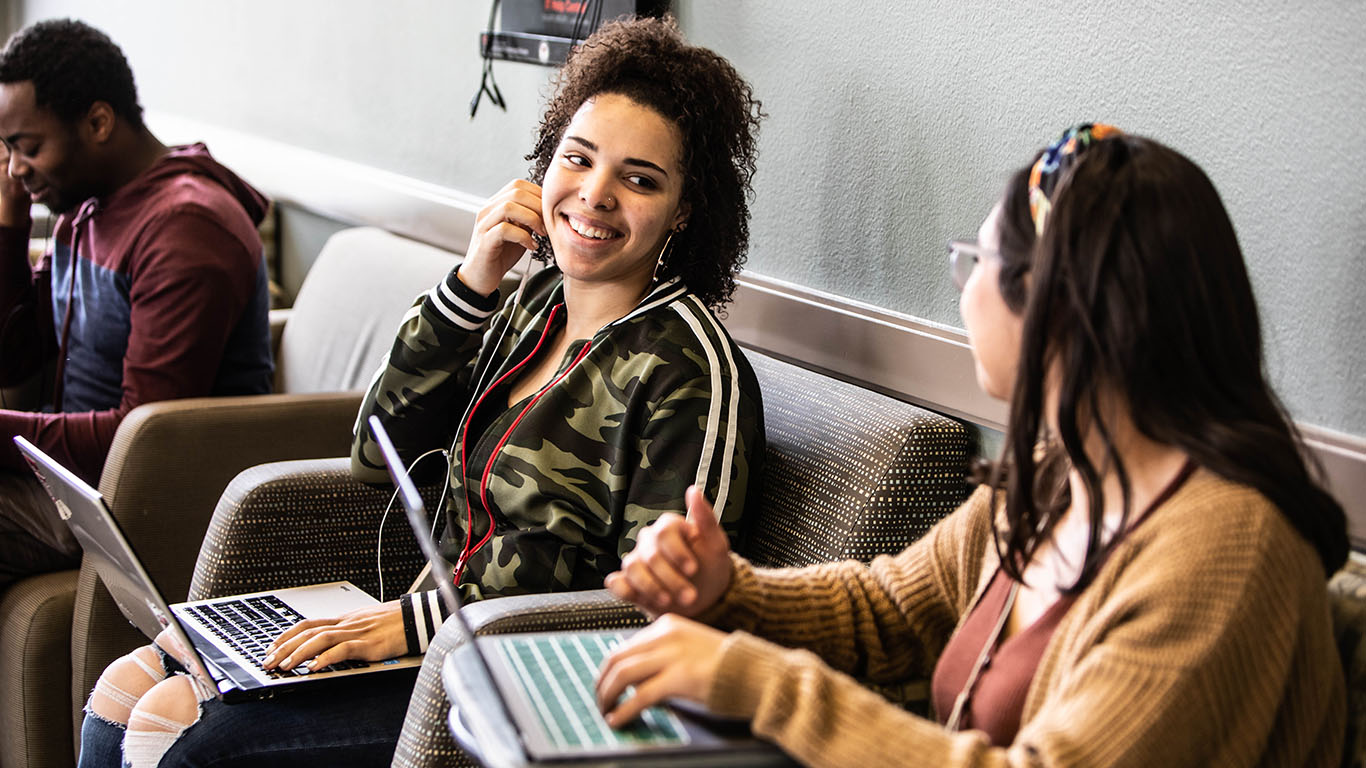 Two students on their laptops in a lounge area.  They are looking at each other and smiling, while one student puts an earbud in her ear.