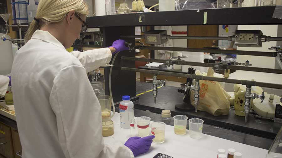 woman in lab gear at counter, performing test with beakers full of liquid