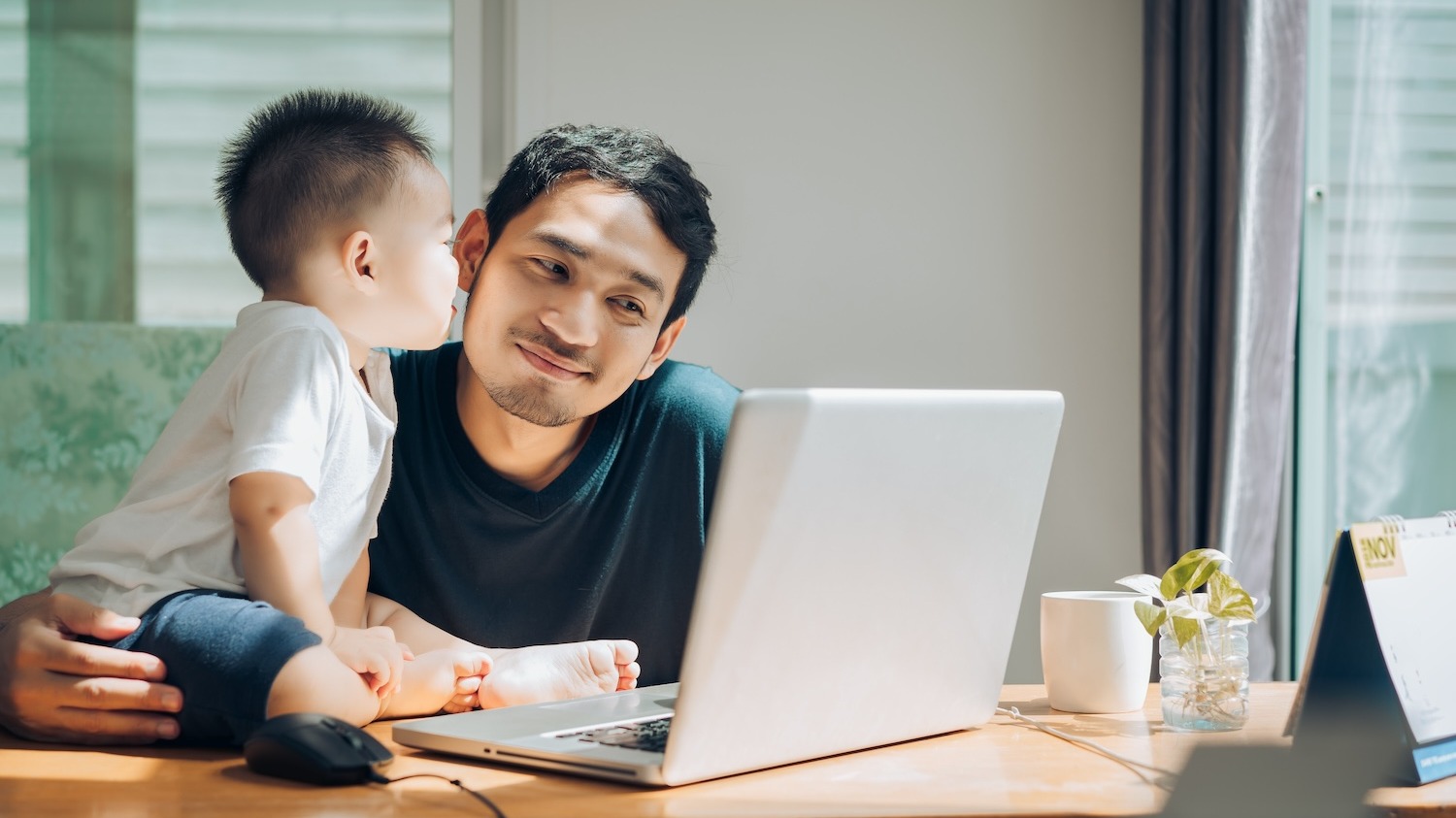 Asian dad receives a kiss from infant while working on laptop