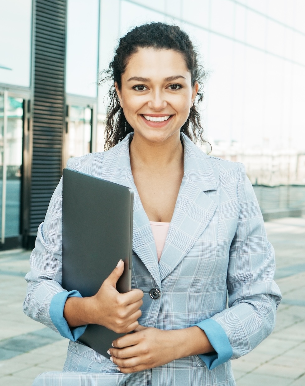 young woman in blazer clutches laptop and smiles at camera
