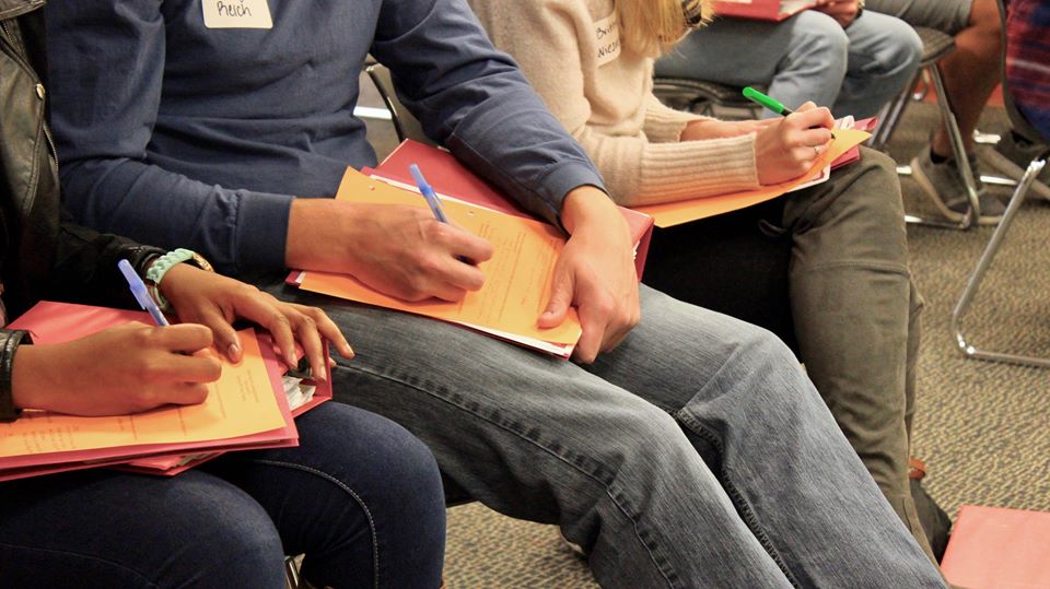 Close-up of a couple students sitting in chairs taking notes.