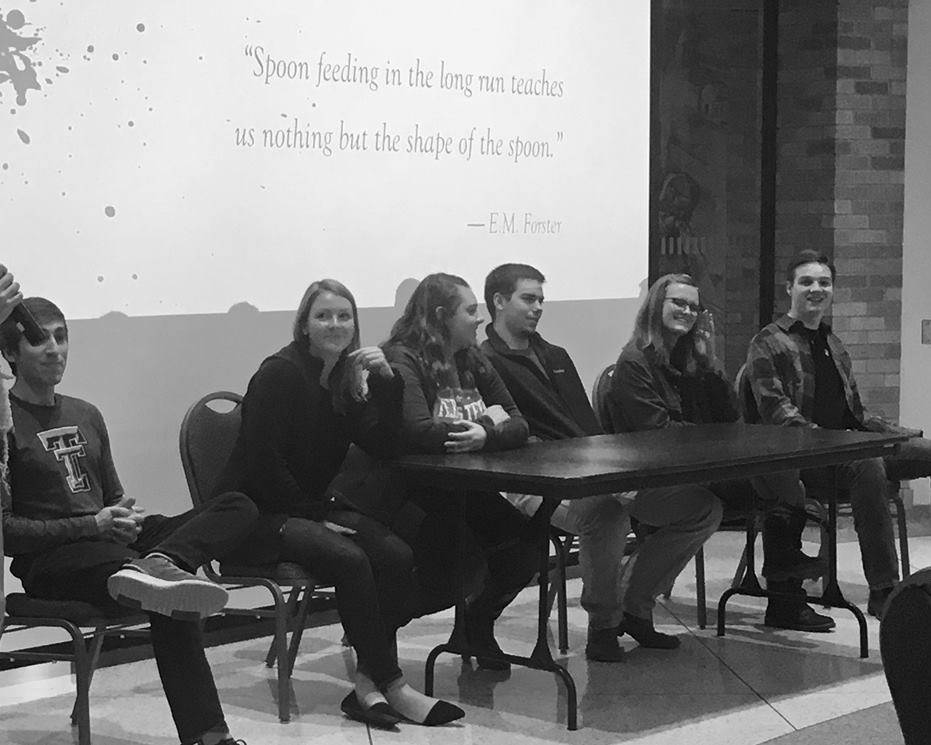 Students sit at table on stage in front of a screen that reads "Spoon feeding in the long run teaches us nothing but the shape of the spoon."